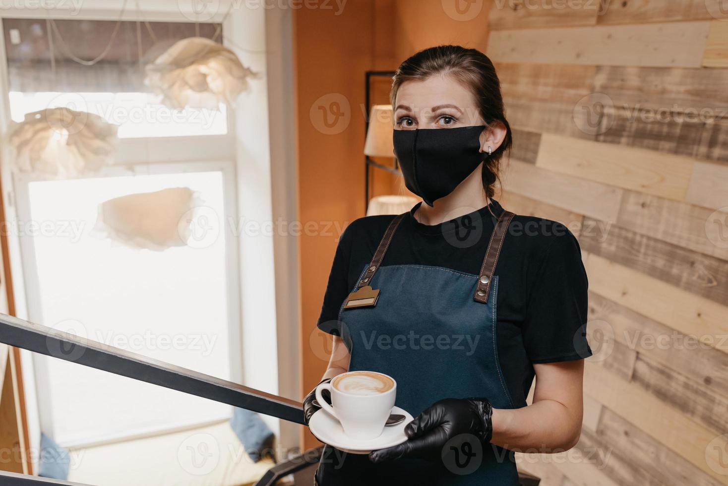 Kellnerin, die eine Gesichtsmaske und Einweghandschuhe trägt, serviert eine Tasse Kaffee foto