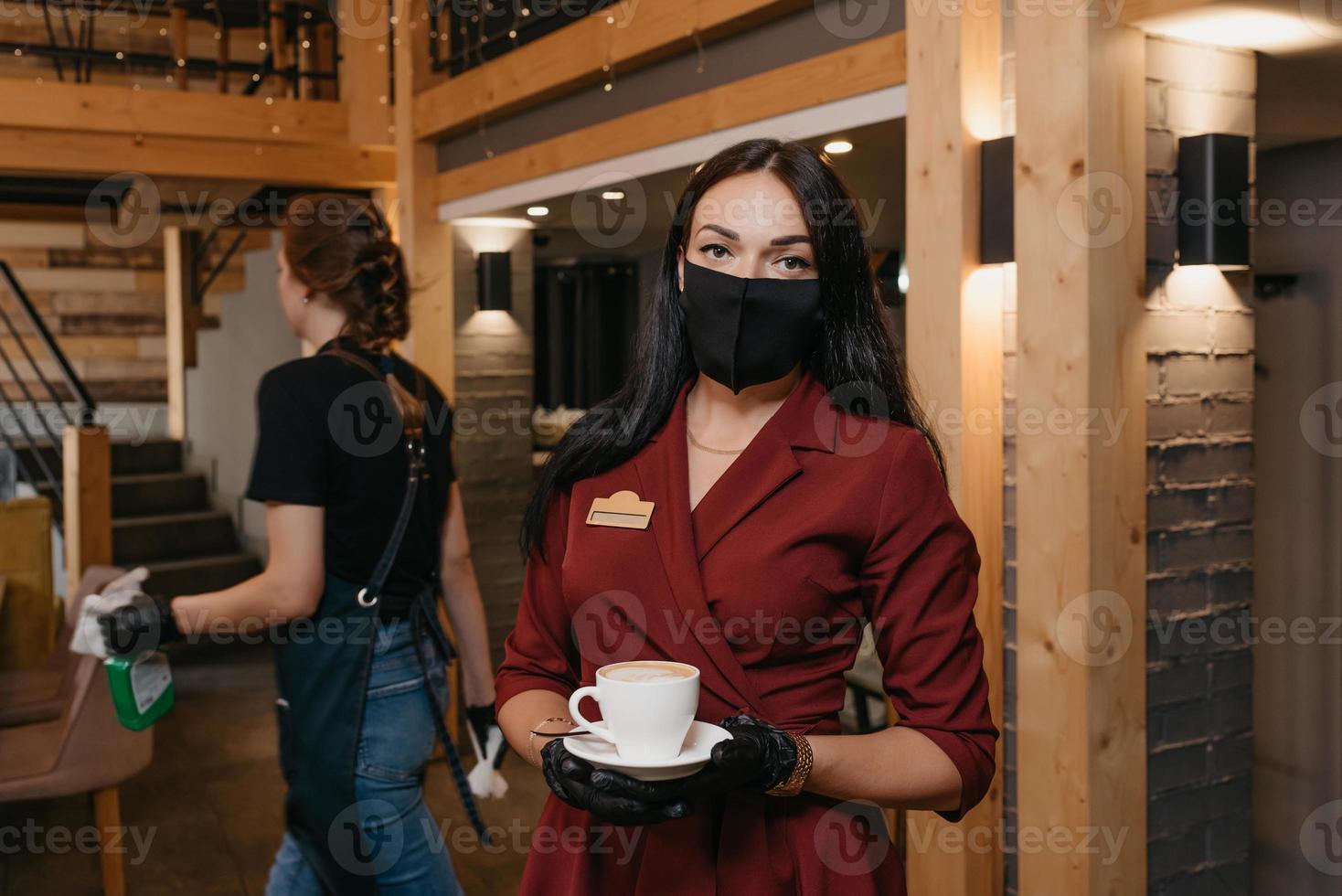 Eine Restaurantmanagerin trägt eine schwarze Gesichtsmaske und Einweghandschuhe mit einer Tasse Kaffee in einem Restaurant foto