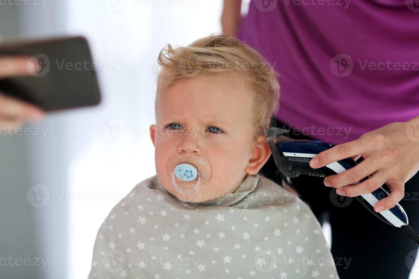 weiblich Hände Schnitt ein Kind mit ein Haar Clipper im ein Friseur. ein Kind Uhren Cartoons auf das Telefon und hält ein Dummy im seine Mund. das zuerst Haarschnitt. foto