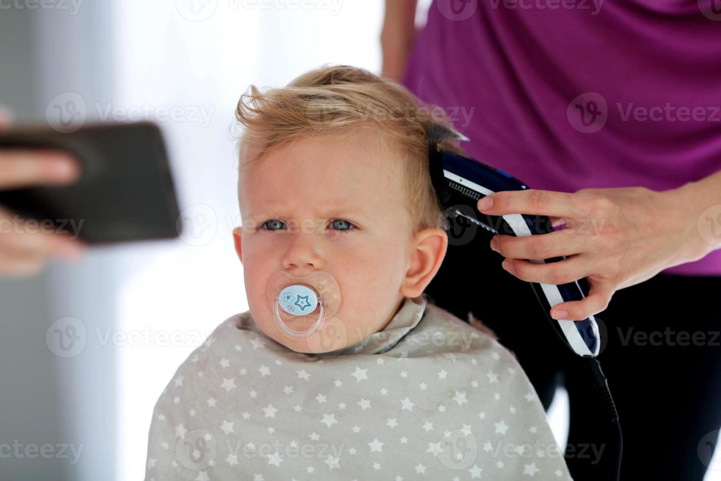 weiblich Hände Schnitt ein Kind mit ein Haar Clipper im ein Friseur. ein Kind Uhren Cartoons auf das Telefon und hält ein Dummy im seine Mund. foto