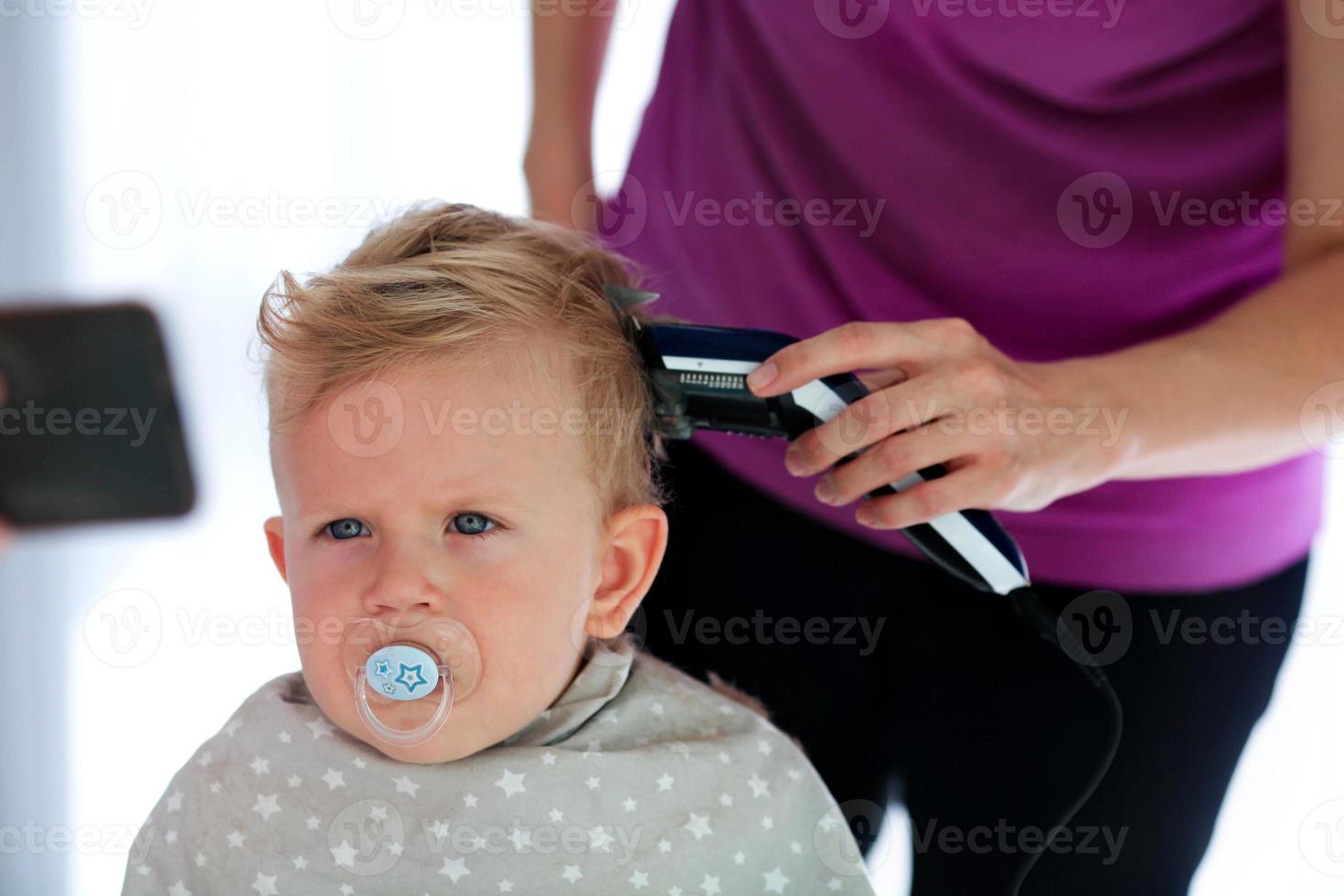 weiblich Hände Schnitt ein Kind mit ein Haar Clipper im ein Friseur. ein Kind Uhren Cartoons auf das Telefon und hält ein Dummy im seine Mund. das zuerst Haarschnitt. foto