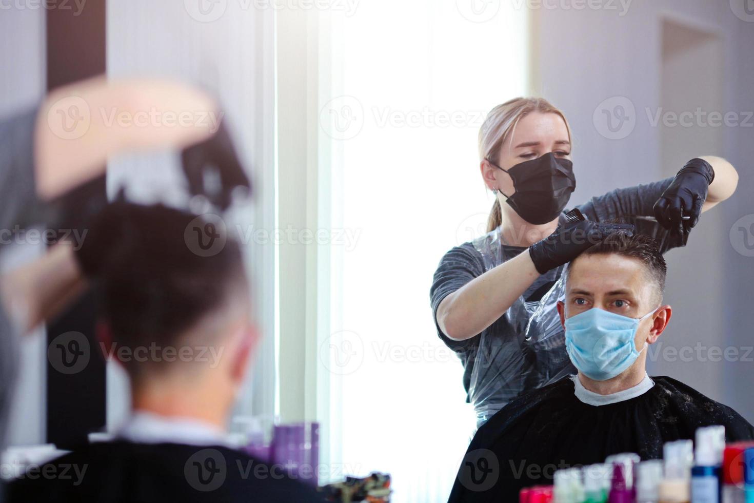 Friseur mit Sicherheit Maße zum COVID-19, schneidet Haar mit ein Schere zu ein Mann im ein Medizin Maske, Sozial Distanz, Schneiden Haar mit ein medizinisch Maske und Gummi Handschuhe foto