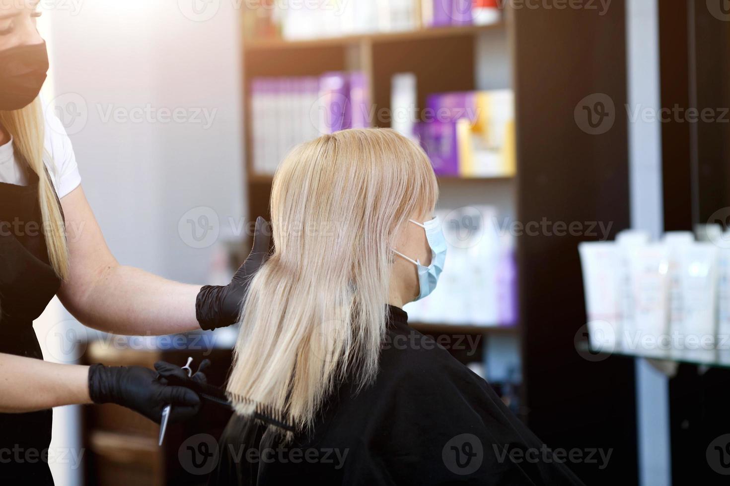 Friseur im Gummi Handschuhe und ein medizinisch Maske Kämmen das Haar von ein Klient. foto