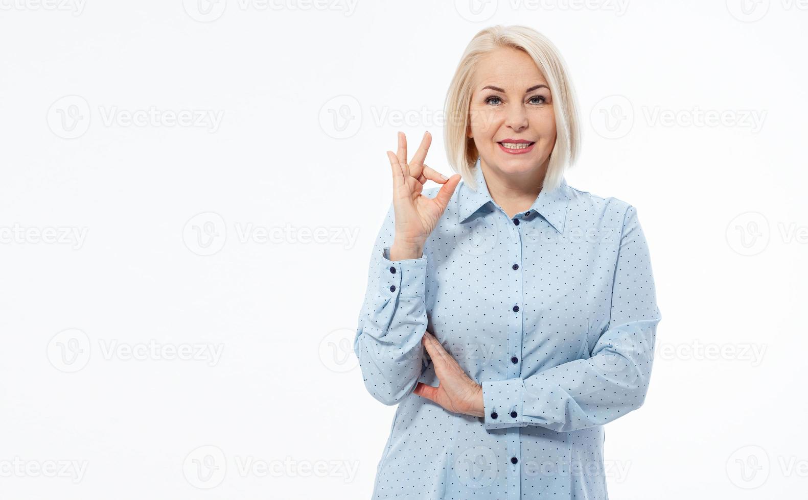 heiter lächelnd Alten grau haarig blond Frau Dame 50er Jahre Jahre alt im Blau Hemd Stehen zeigen in Ordnung Geste suchen Kamera isoliert auf Weiß Hintergrund. foto