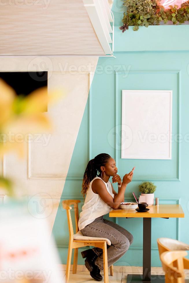 jung schwarz Frau mit Handy, Mobiltelefon Telefon während haben Frühstück im das Cafe foto