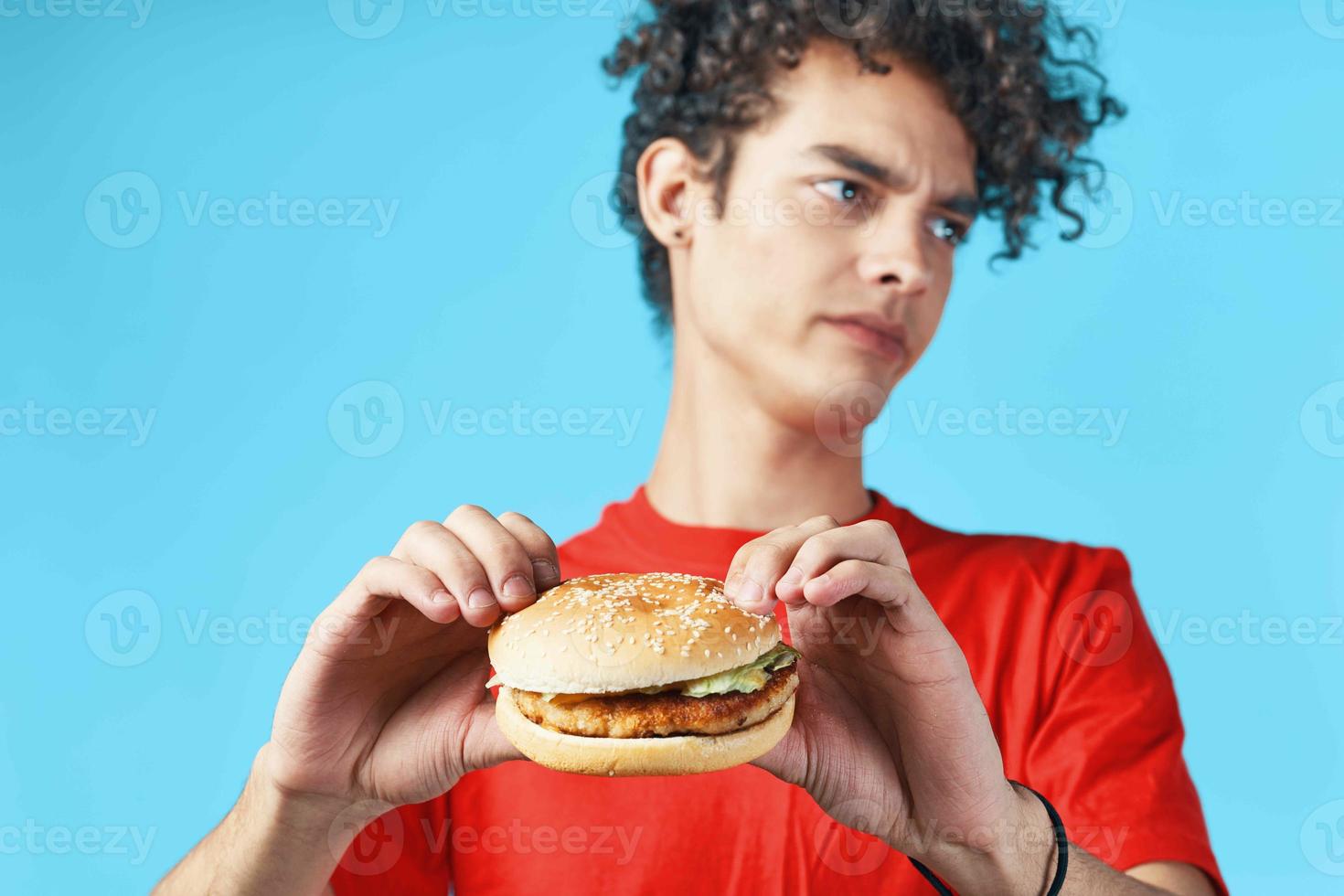 heiter Kerl mit lockig Haar im ein rot T-Shirt mit ein Hamburger im seine Hände schnell Essen Diät foto