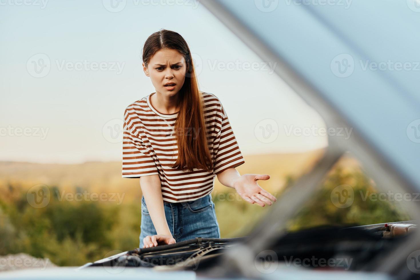 ein traurig Frau hat geöffnet das Kapuze von ein gebrochen Nieder Auto und ist suchen zum das Ursache von das Nervenzusammenbruch auf ein Straße Ausflug allein foto
