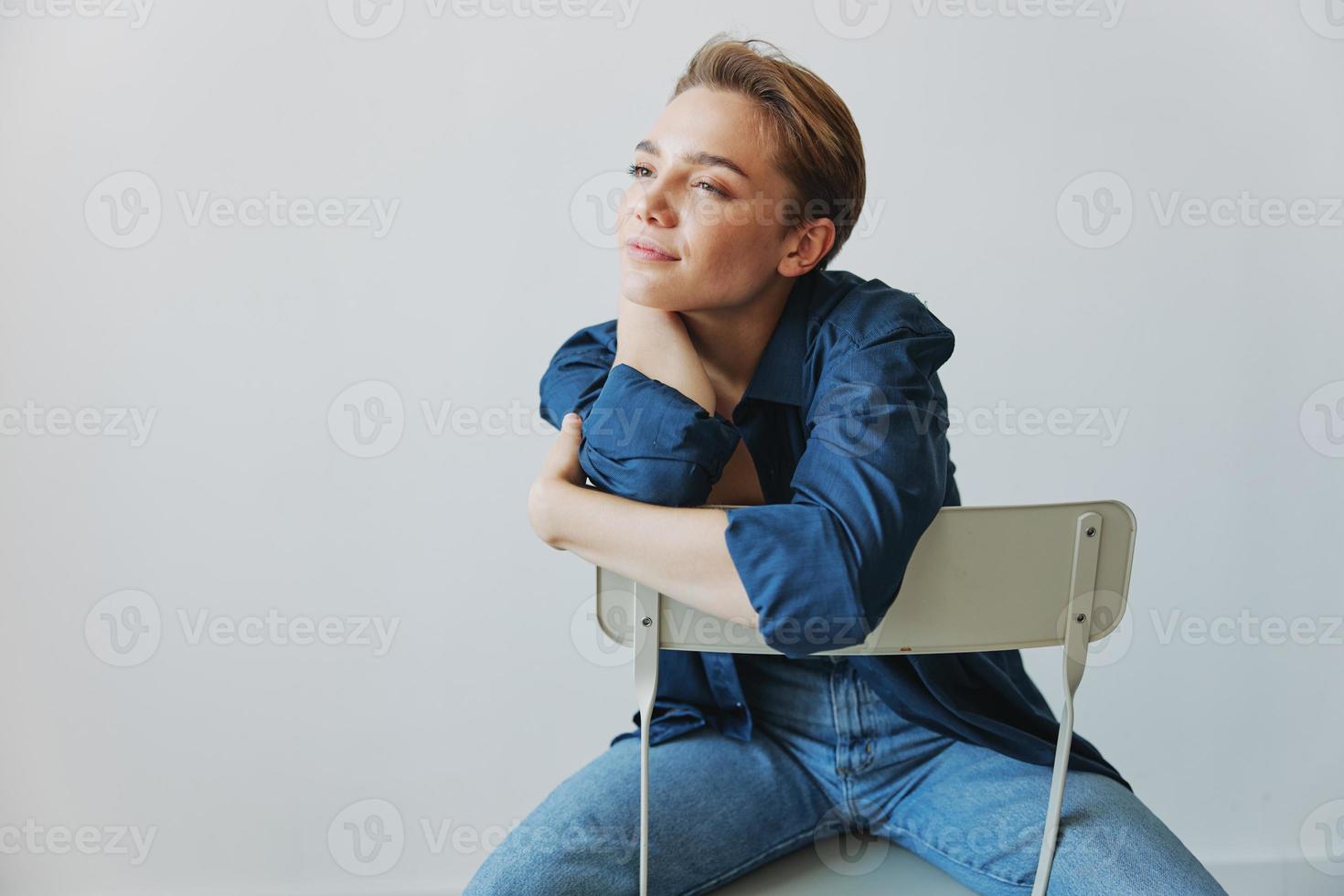 ein jung Frau Sitzung im ein Stuhl beim Zuhause lächelnd mit Zähne mit ein kurz Haarschnitt im Jeans und ein Denim Hemd auf ein Weiß Hintergrund. Mädchen natürlich posiert mit Nein Filter foto