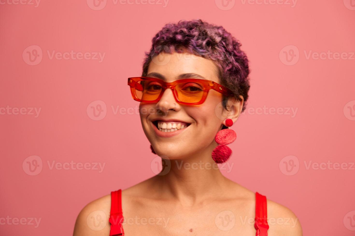 Mode Porträt von ein Frau mit ein kurz Haarschnitt im farbig Sonnenbrille mit ungewöhnlich Zubehör mit Ohrringe lächelnd auf ein Rosa hell Hintergrund foto