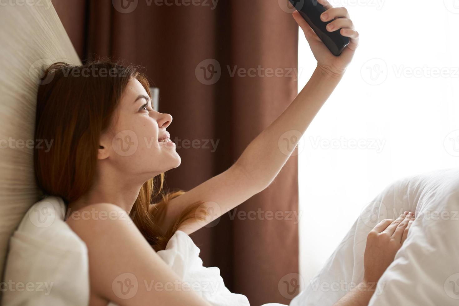 Frau mit Telefon im Hand im Bett in der Nähe von das Fenster, Vorhang foto