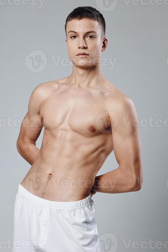 Athlet mit ein aufgepumpt Torso auf ein grau Hintergrund hält seine Hände hinter seine zurück und Weiß kurze Hose foto