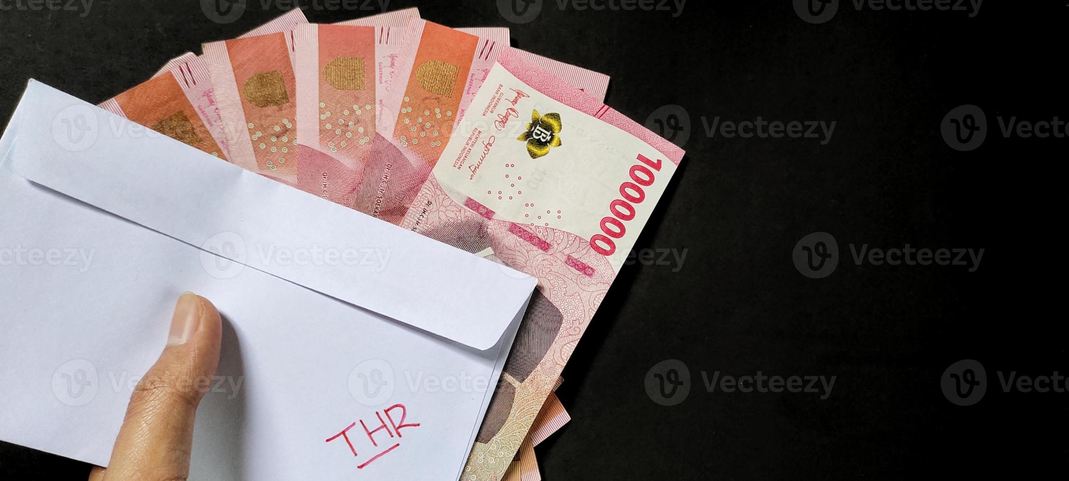 Mann halten ein Weiß Briefumschlag geschrieben von thr und Neu indonesisch Banknoten, in der Regel Tunjangan Hari raya oder namens thr sind gegeben zu Angestellte voraus von eid. isoliert auf schwarz Hintergrund und oben Aussicht foto