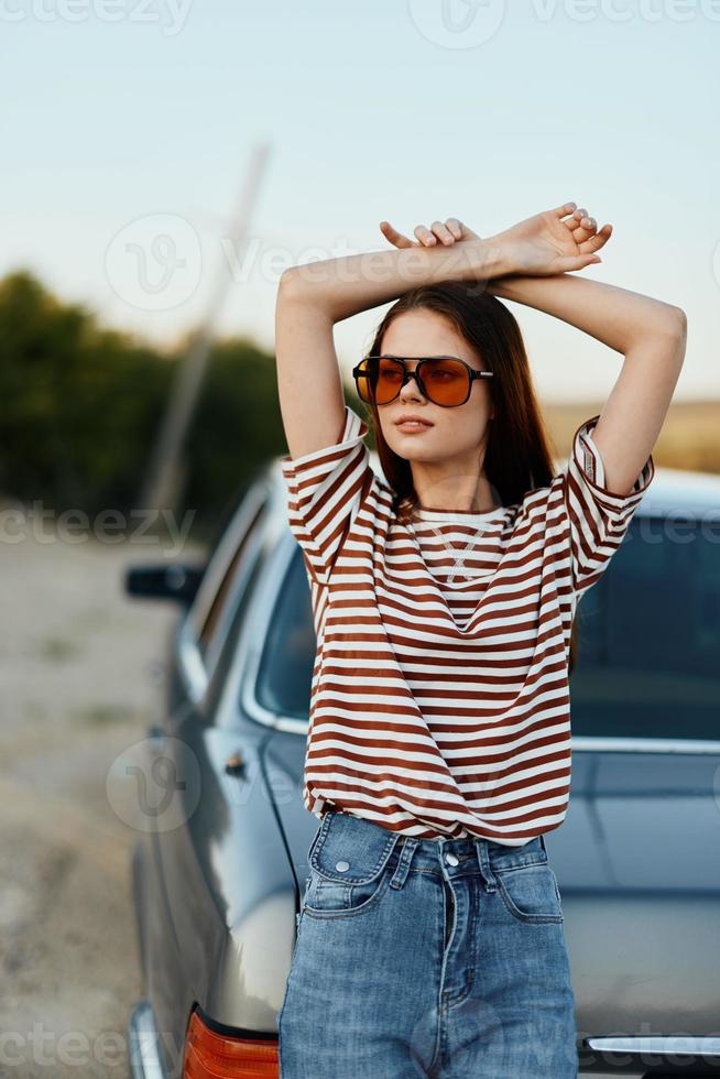 stilvoll Frau im gestreift T-Shirt Sonnenbrille und Jeans in der Nähe von grau Auto im Natur foto