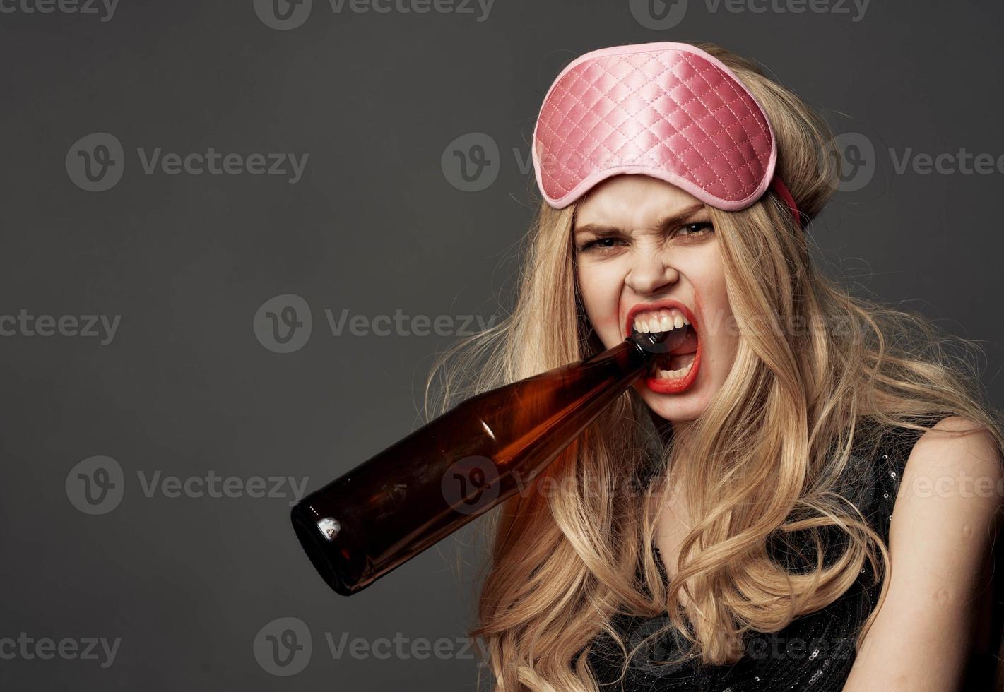 wütend Frau mit ein Flasche von Bier im ihr Hand mit hell bilden und ein Rosa Schlaf Maske foto