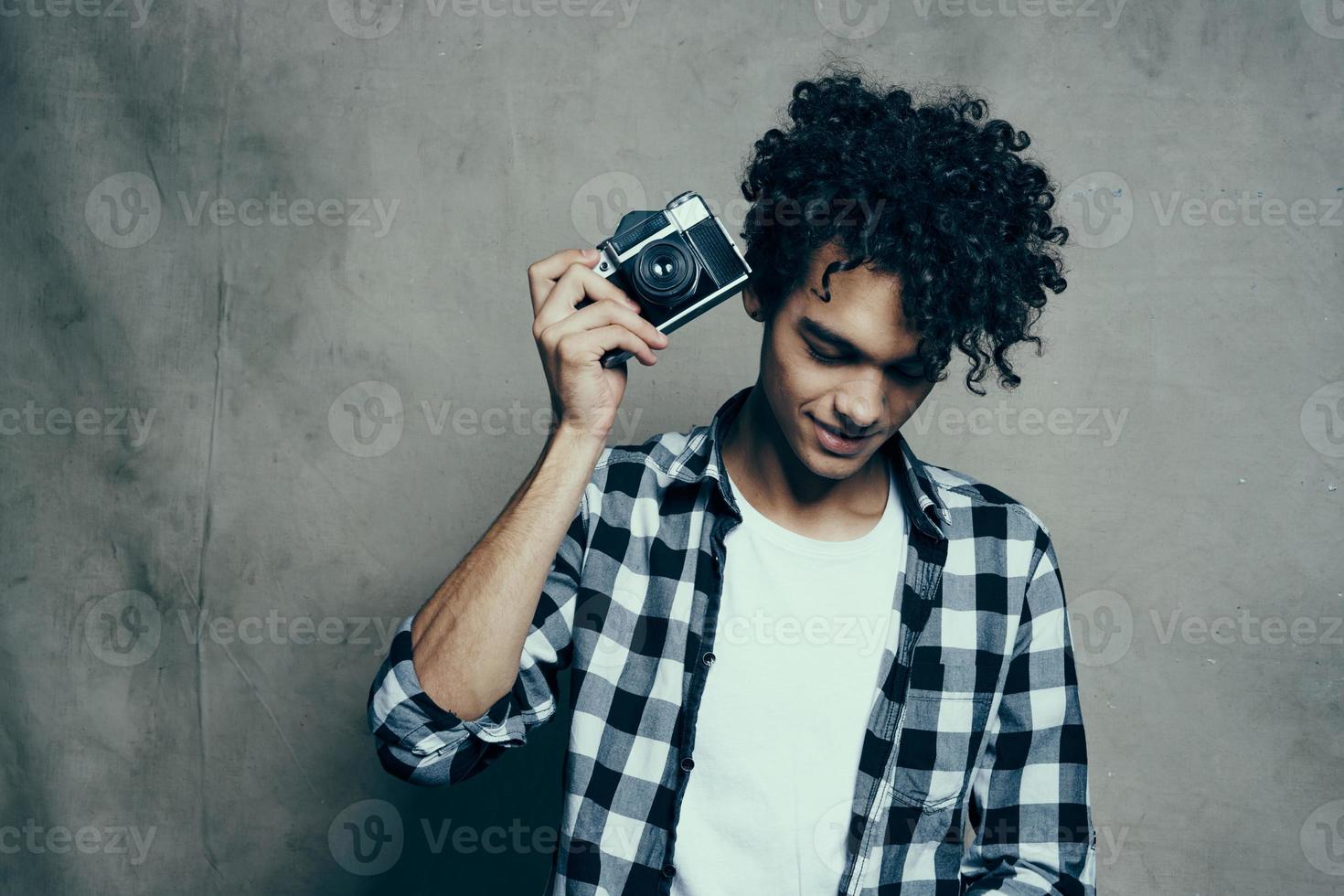 jung Mann mit ein Kamera im Hand und im ein Plaid T-Shirt auf ein grau Hintergrund drinnen Fotograf foto