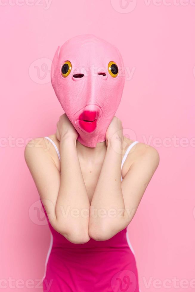 Frau Stehen im ein Rosa Fisch Maske auf ihr Kopf, ein provokativ verrückt Halloween sehen, Rosa Hintergrund im das Studio foto