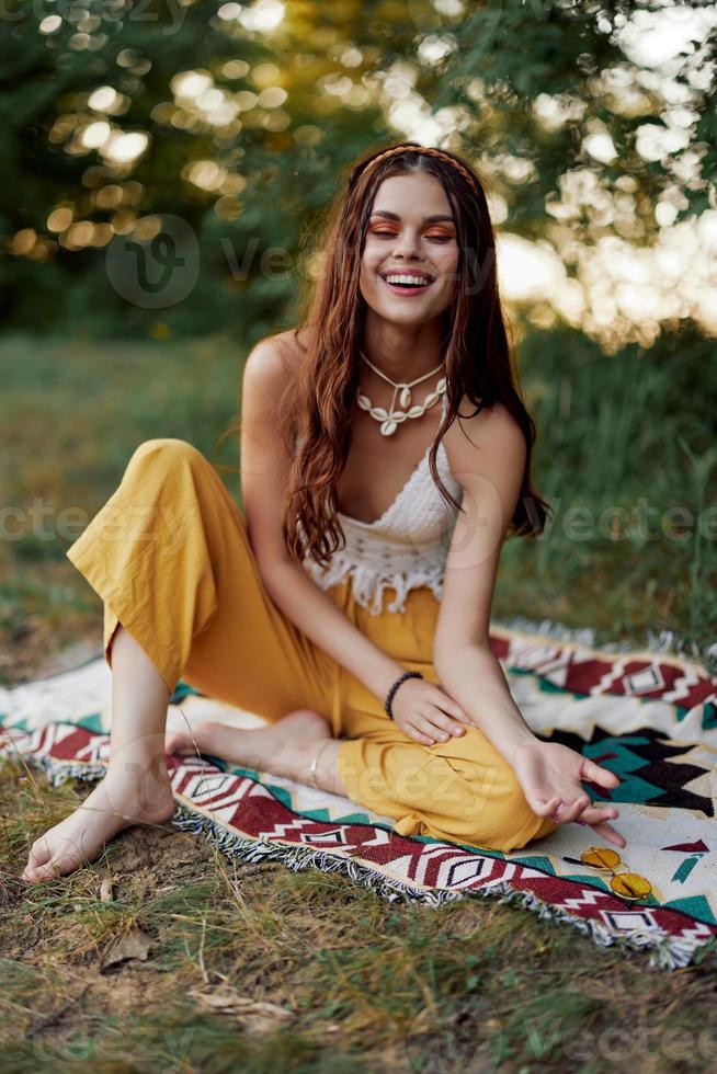 ein jung schön Hippie Frau sitzt auf das Boden im das fallen im Öko Kleidung im das Sonnenuntergang Licht und lächelt Mit freundlichen Grüßen zum das Kamera foto