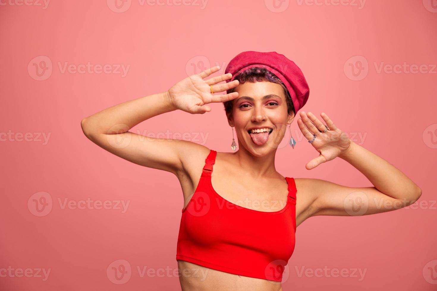 jung sportlich Frau mit ein kurz Haarschnitt und lila Haar im ein rot oben und ein Rosa Hut mit ein sportlich Zahl lächelt und Grimassen suchen beim das Kamera auf ein Rosa Hintergrund foto