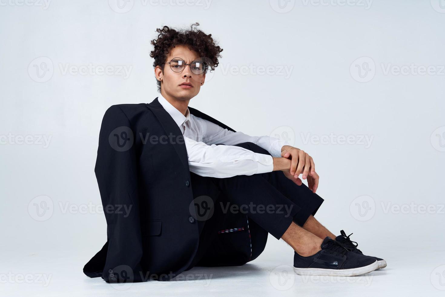 modisch Kerl mit lockig Haar im ein klassisch passen und Turnschuhe auf das Fußboden drinnen Fotografie Studio foto