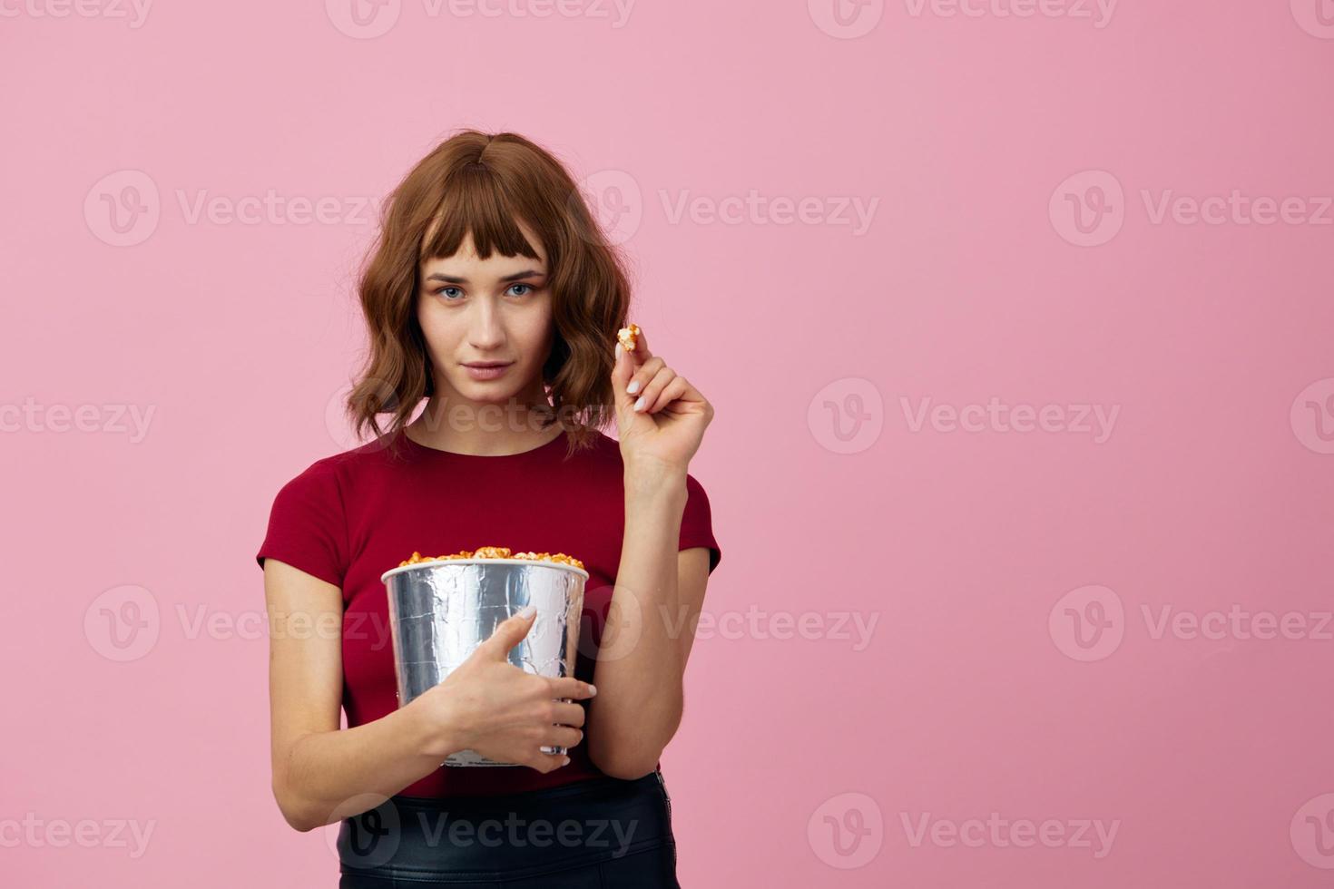 aufgeregt verwirrt süß Rothaarige Dame im rot T-Shirt mit Popcorn bereit zum Film Abend posieren isoliert auf Über Rosa Studio Hintergrund. Kopieren Raum Banner. Mode Kino Konzept. Unterhaltung Angebot foto