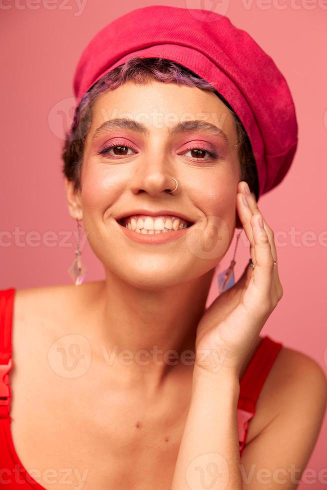 jung sportlich Frau mit ein kurz Haarschnitt und lila Haar im ein rot oben und ein Rosa Hut mit ein sportlich Zahl lächelt und Grimassen suchen beim das Kamera auf ein Rosa Hintergrund foto