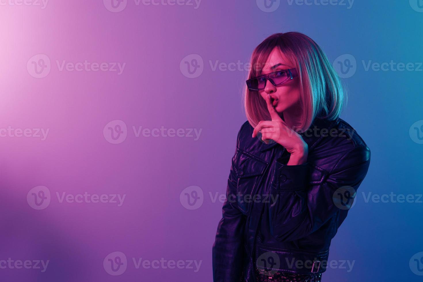 süß schön blond Frau im Leder Jacke prickelnd Kleid modisch Sonnenbrille sagen Sein ruhig mit Finger auf Lippen shhh Geste posieren isoliert im Blau Rosa Farbe Licht Hintergrund. Neon- Party Konzept foto