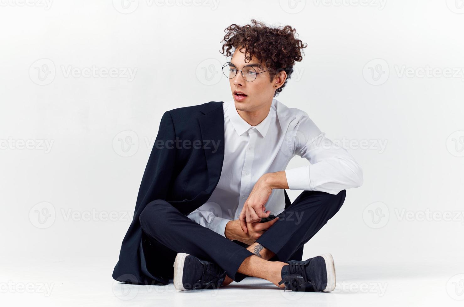 lockiges Haar Kerl im modisch Kleider und Turnschuhe sitzt auf das Fußboden im ein hell Zimmer im voll Wachstum foto