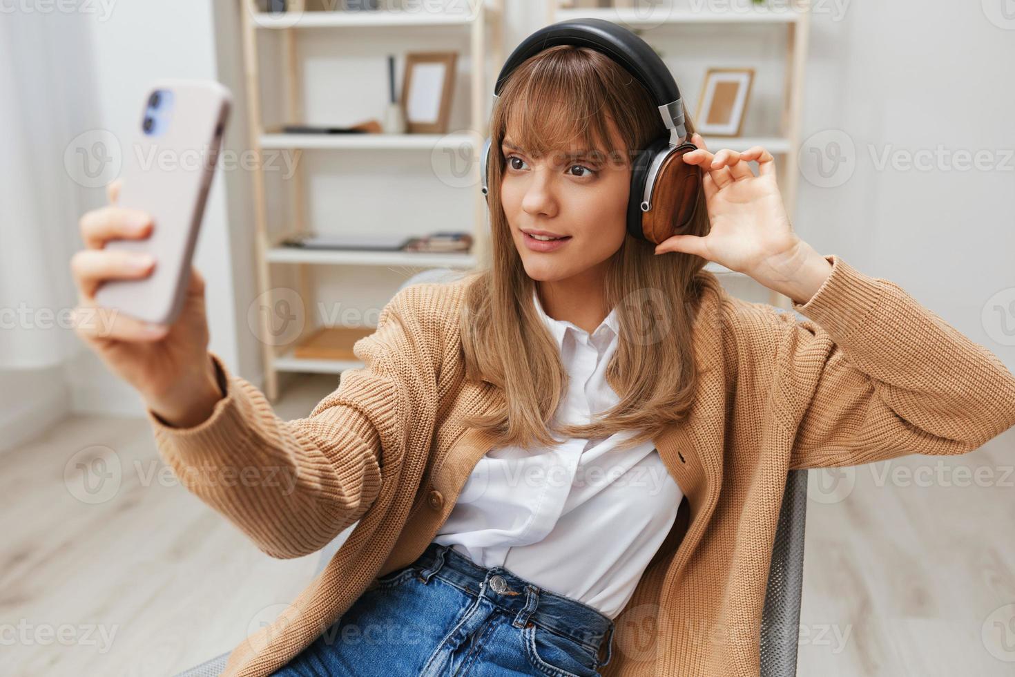 froh jung blond Dame im warm Sweatshirt im Kopfhörer Hör mal zu Favorit Lieder tun Selfie Video Anruf Sitzung im Sessel beim modern Zuhause Innere. Musik- Zeit entspannend cool Wiedergabeliste Konzept. Kopieren Raum foto