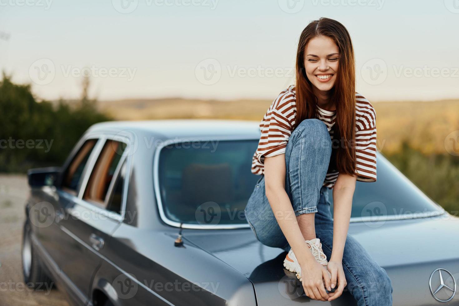 ein jung Frau sitzt auf das Kofferraum von ein Auto, lacht und ruht nach ein schwierig Straße und bewundert Natur mit ein schön Sicht. anhalten ist ebenfalls Teil von das Reise foto
