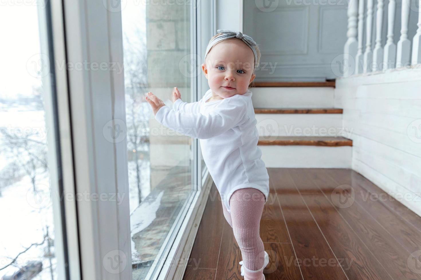 Kleines krabbelndes Mädchen, ein Jahr alt, sitzt lächelnd und lachend auf dem Boden im hellen Wohnzimmer in der Nähe des Fensters foto