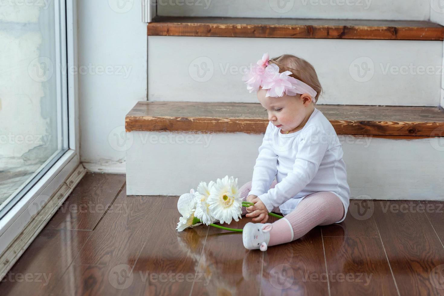wenig Baby Mädchen tragen Frühling Kranz Standort auf Fußboden im hell Licht Leben Zimmer in der Nähe von Fenster und spielen mit Gerbera Blumen foto