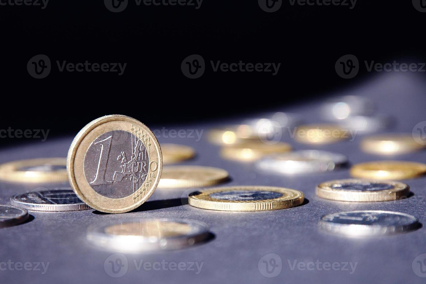 einer Euro Münze Stehen auf Kante leuchtenden im Scheinwerfer gegen viele anders Münzen auf dunkel Hintergrund. Geld fließen foto