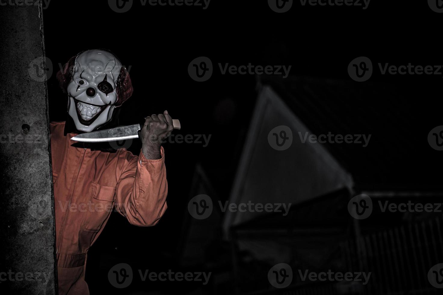 asiatisch gut aussehend Mann tragen Clown Maske mit Waffe beim das Nacht Szene, Halloween Festival Konzept, Schrecken unheimlich Foto von ein Mörder im Orange Stoff
