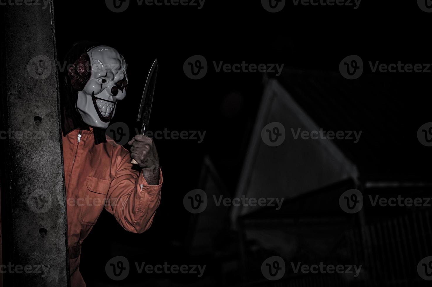 asiatisch gut aussehend Mann tragen Clown Maske mit Waffe beim das Nacht Szene, Halloween Festival Konzept, Schrecken unheimlich Foto von ein Mörder im Orange Stoff