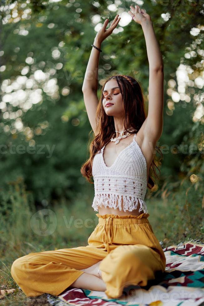 ein jung Hippie Frau meditiert im Natur im das Park, Sitzung im ein Lotus Position auf ihr bunt Plaid und genießen Harmonie mit das Welt im Öko-Kleidung im das Herbst foto