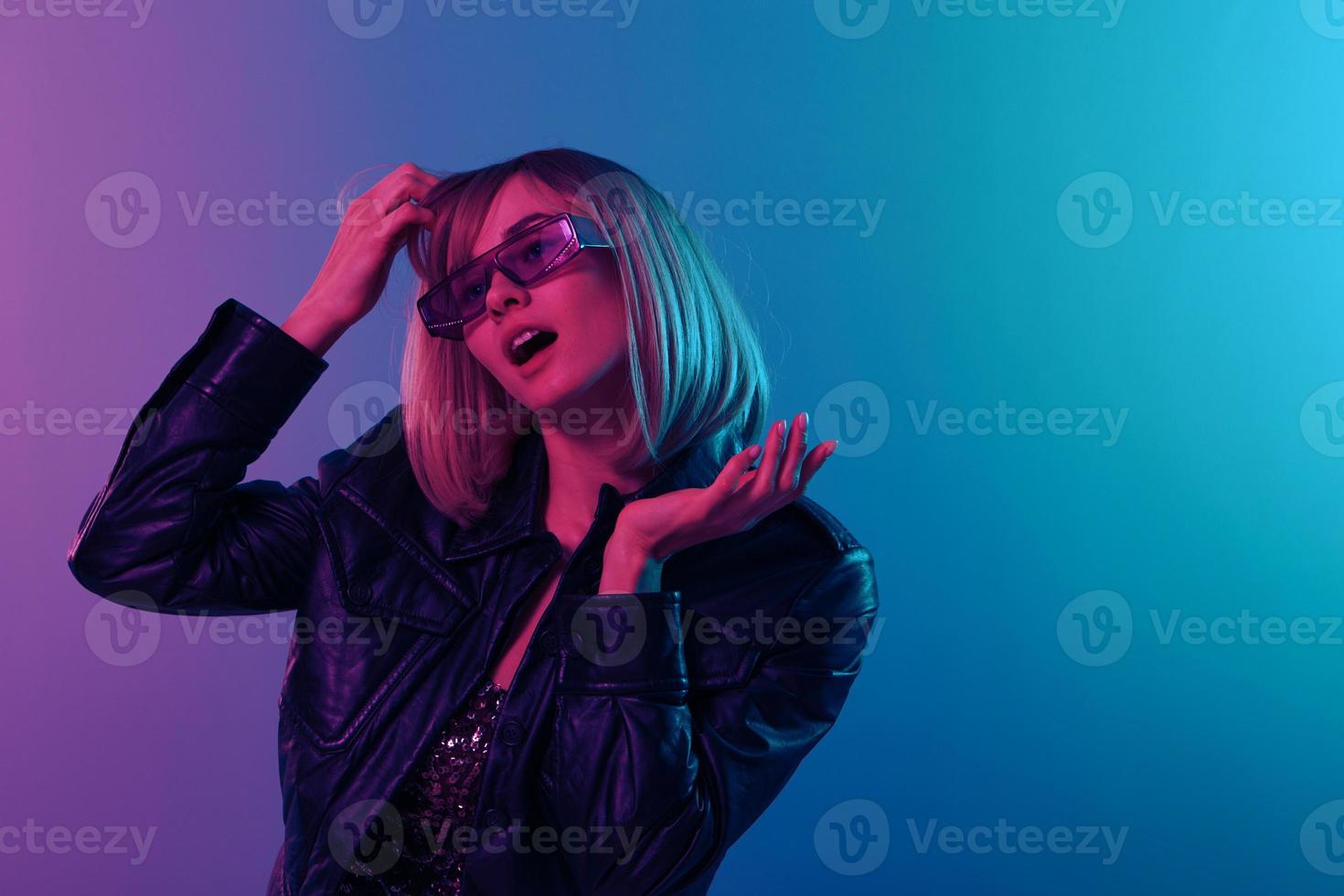 sexy schön blond Frau im Leder Jacke prickelnd Kleid modisch Sonnenbrille berühren Haar aussehen beiseite posieren isoliert im Farbe Licht Studio Mauer Hintergrund. Neon- Party Cyberpunk Konzept. Kopieren Raum foto