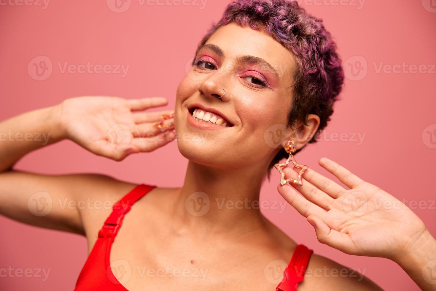 Mode Porträt von ein Frau mit ein kurz Haarschnitt von lila Farbe und ein Lächeln mit Zähne im ein rot oben auf ein Rosa Hintergrund Glück foto