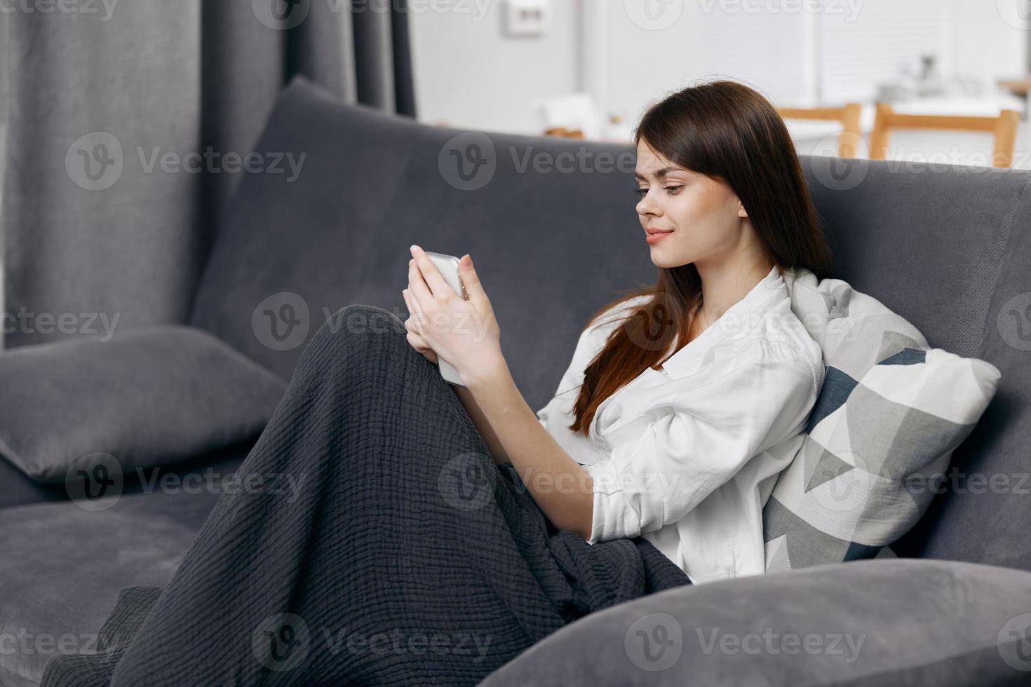 Frau Sitzung auf das Couch mit ein Decke auf ihr Runde und ein Handy, Mobiltelefon Telefon im ihr Hand foto