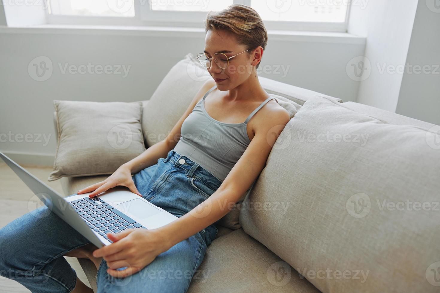 Teenager Mädchen Freiberufler mit Laptop Sitzung auf Couch beim Zuhause lächelnd im Zuhause Kleider und Brille mit kurz Haarschnitt, Lebensstil mit Nein Filter, kostenlos Kopieren Raum foto