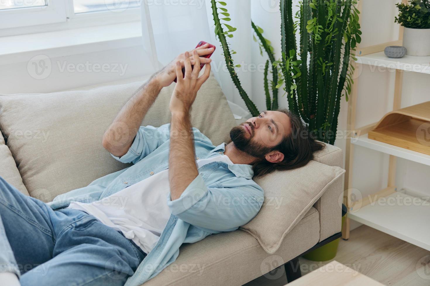 ein Mann mit ein Bart Lügen auf das Couch während das Tag beim Zuhause und sieht aus beim seine Telefon entspannend auf seine Tag aus, ein Mann Glücksspiel auf das Lager Markt online auf seine Telefon foto