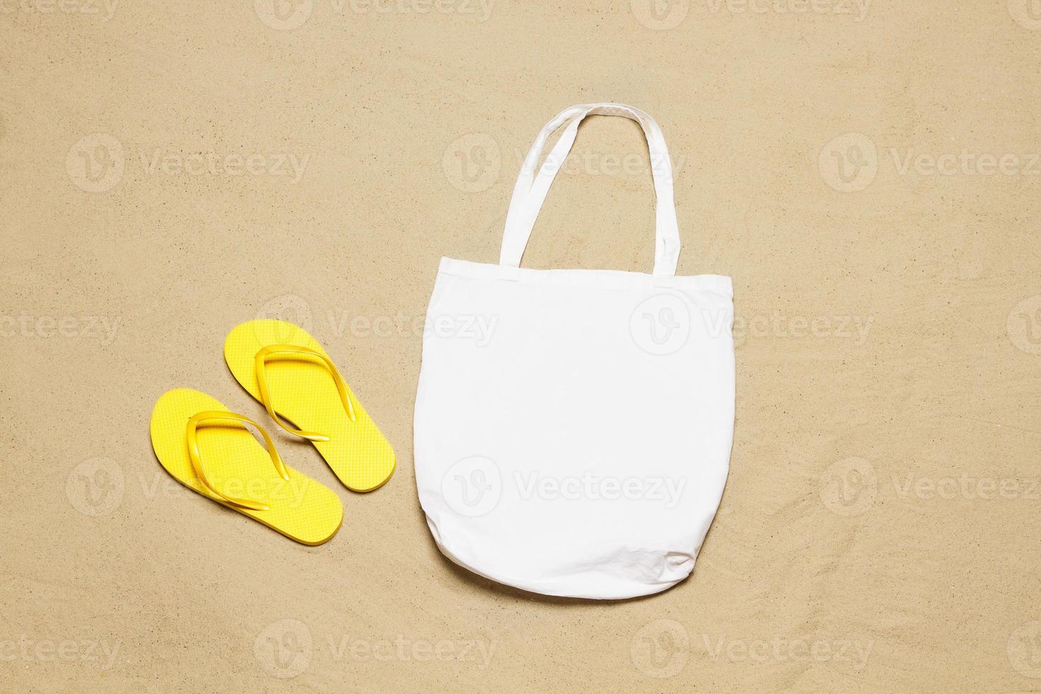 Attrappe, Lehrmodell, Simulation Weiß Käufer Handtasche Strand Sand Hintergrund. oben Aussicht Kopieren Raum Einkaufen Öko wiederverwendbar Tasche. Flip Flops Zubehör. Vorlage leer oben Aussicht Baumwolle Material Segeltuch Tuch. leeren Attrappe, Lehrmodell, Simulation Strand foto