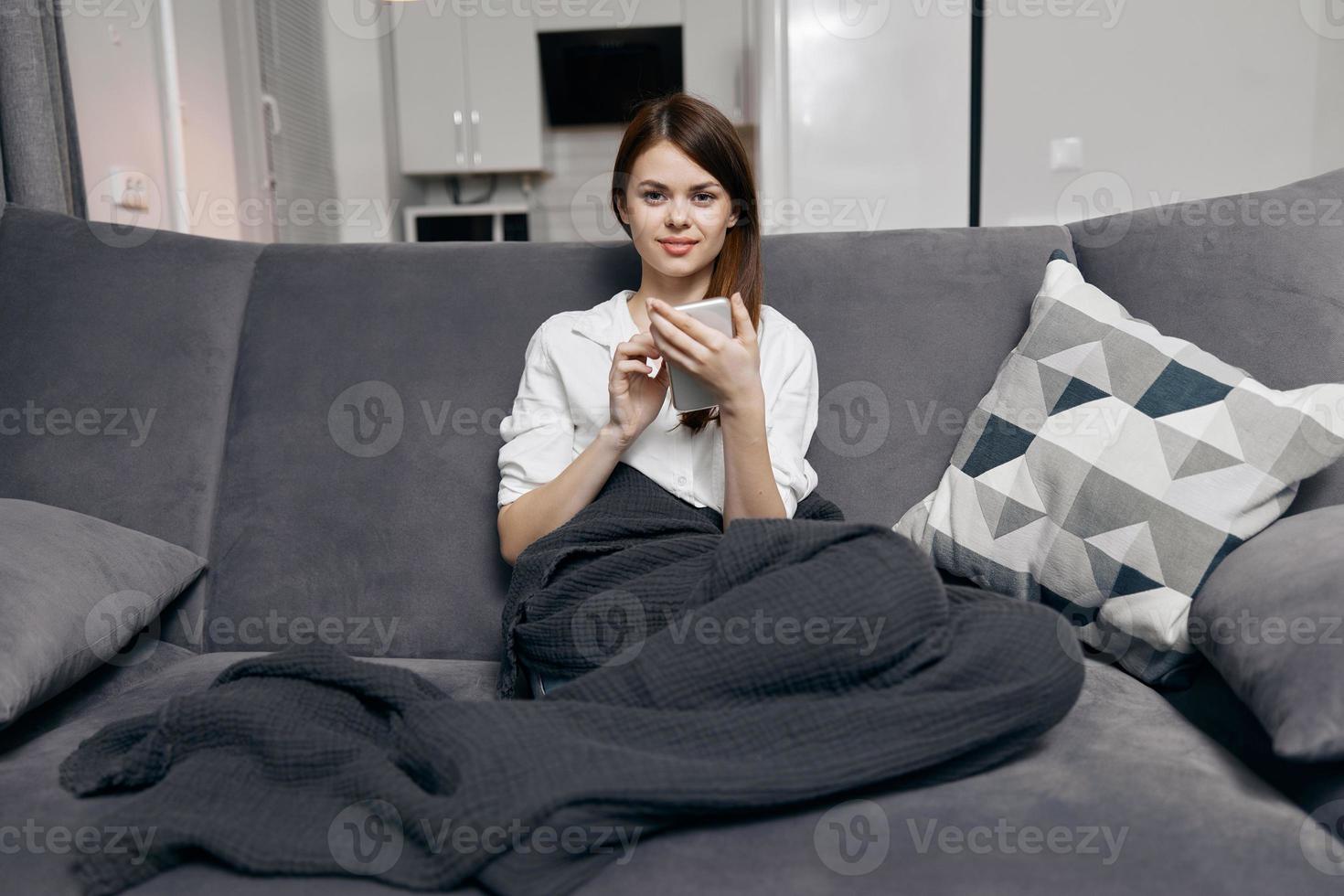 ein Frau im ein Wohnung mit ein Handy, Mobiltelefon Telefon sitzt auf ein Sofa bedeckt mit ein Decke foto