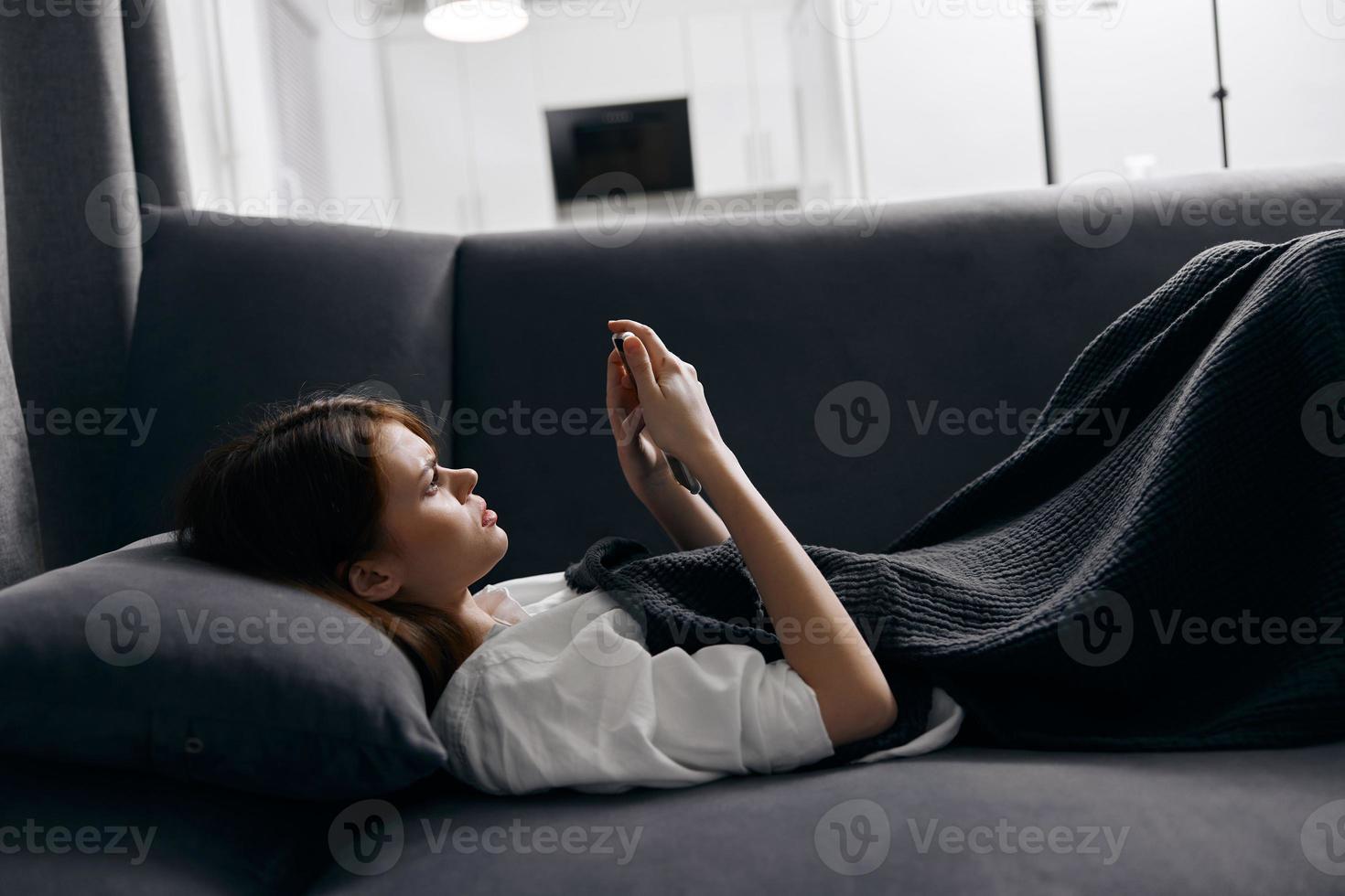 ein Frau Lügen auf ein Sanft Sofa drinnen und sieht aus beim ein Handy, Mobiltelefon Telefon Bildschirm foto