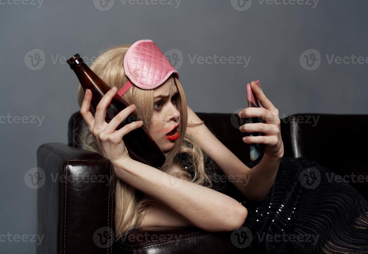 ein betrunken Frau mit ein Flasche von Bier sieht aus beim ein Handy, Mobiltelefon Telefon im ihr Hand und Lügen auf das Couch foto