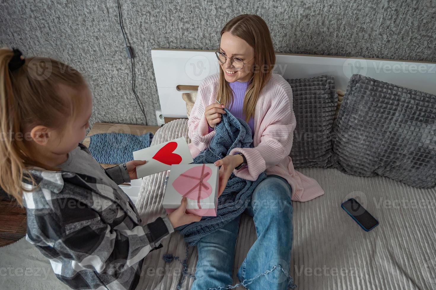 das Tochter gratuliert ihr Mutter auf Mutter Tag, gibt ihr ein Box mit ein Geschenk und ein Glückwunsch Öffner mit ein Herz gemacht durch Sie selber. ein Frau strickt im ihr Schlafzimmer. foto