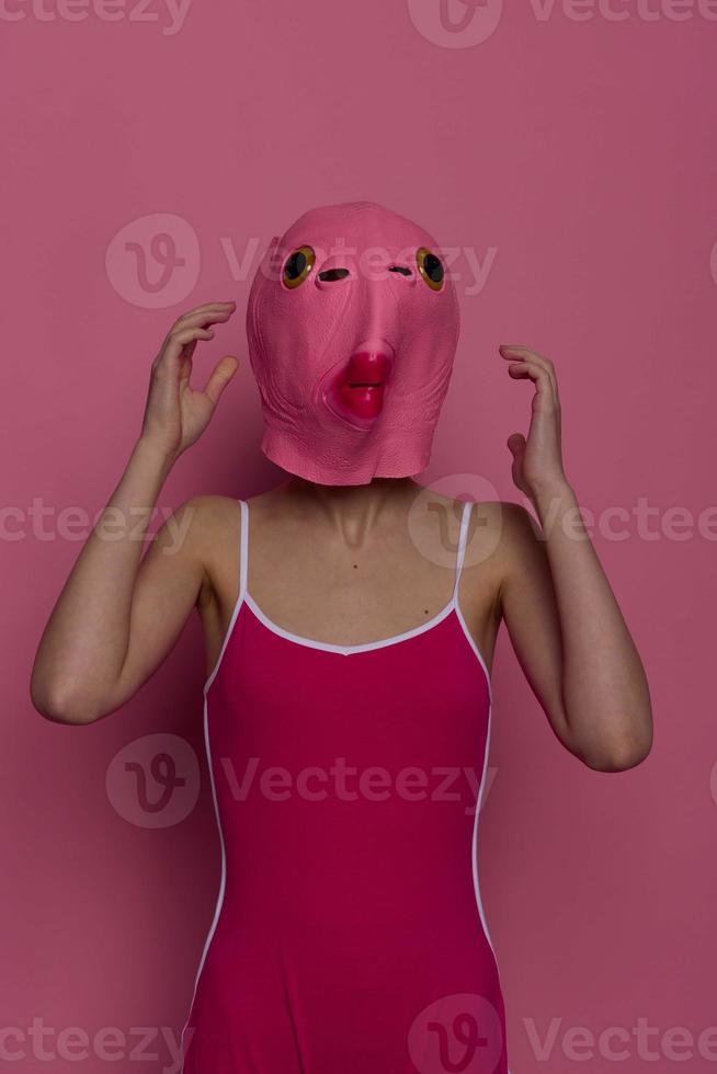 Frau Stehen im ein Rosa Fisch Maske auf ihr Kopf, ein provokativ verrückt Halloween sehen, Rosa Hintergrund im das Studio foto