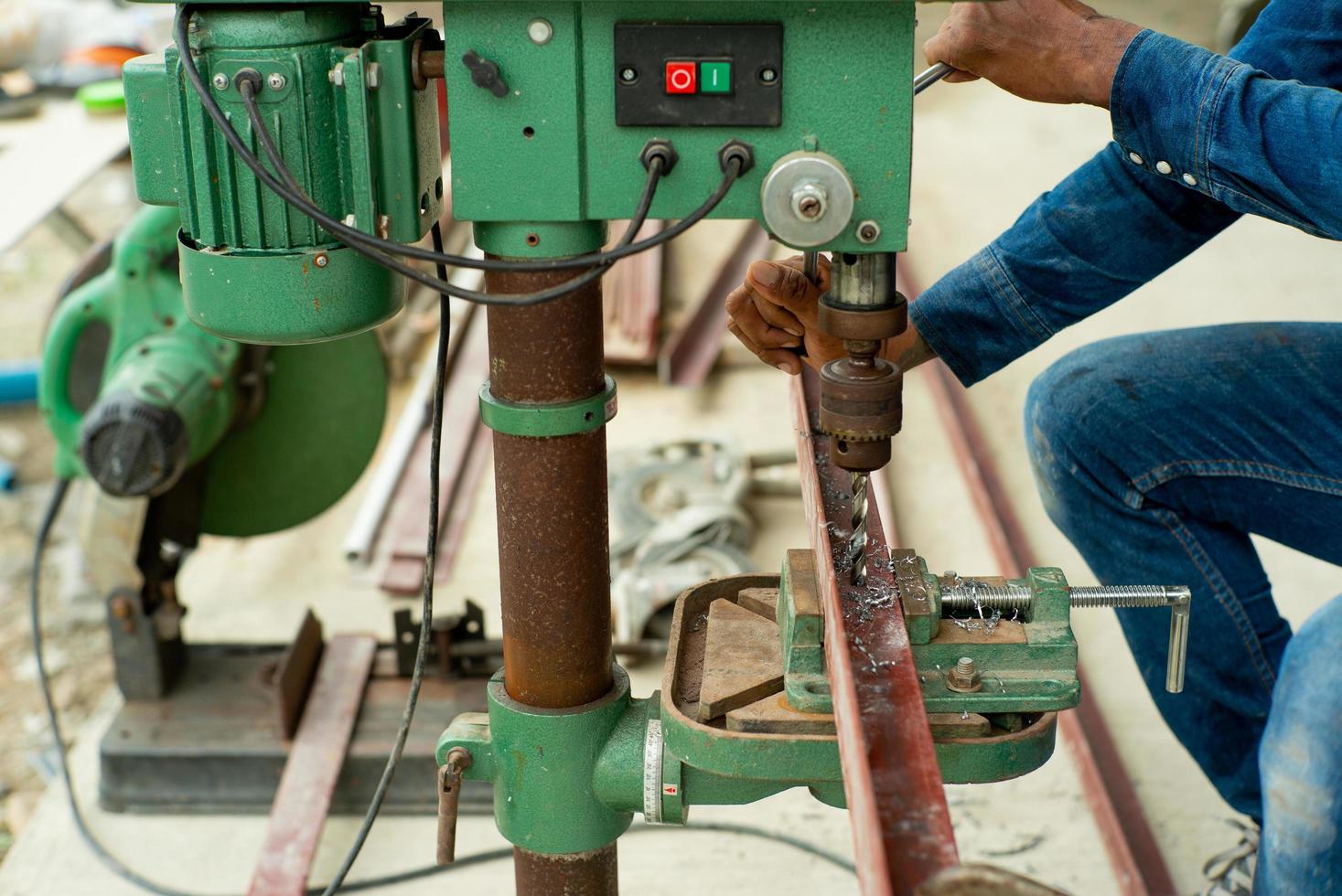 Der selektive Fokus der Nahaufnahme auf die Hände des Arbeiters steuert die elektrische Bohrmaschine zum Bohren des Lochs in der Winkelstahlstange auf der Baustelle foto