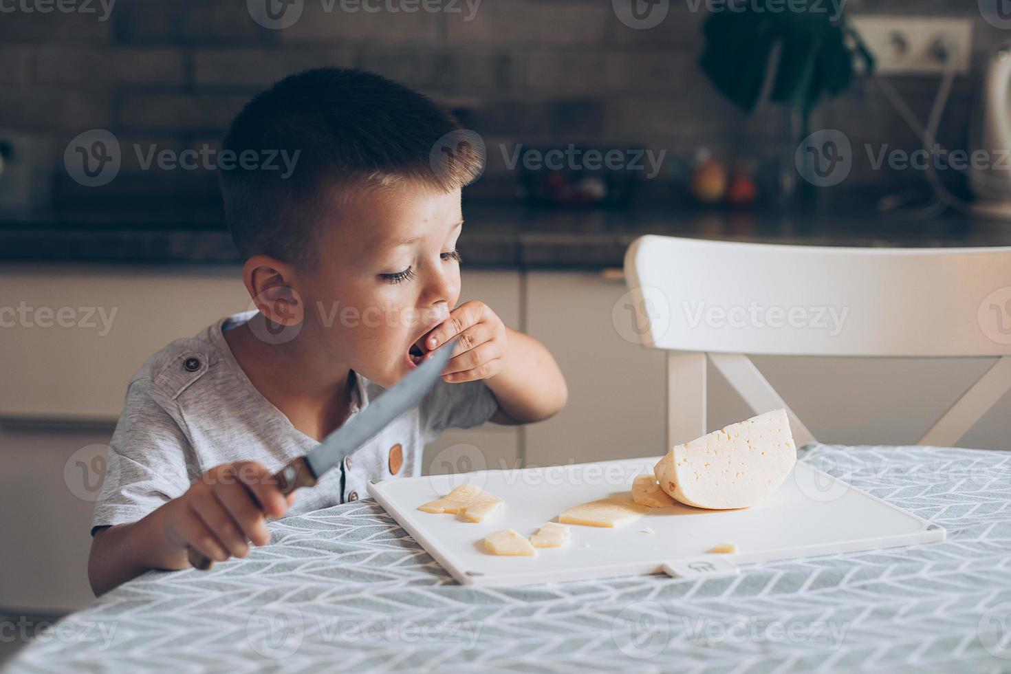 süß Junge 4-5 Jahre alt mit Messer Schneiden ein Käse auf das Schneiden Tafel auf das Tabelle im das Küche. foto