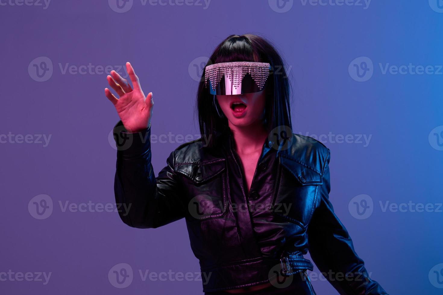 verwirrt ziemlich genial brünett Dame im Leder Jacke modisch spiegelnd Sonnenbrille berühren unsichtbar Objekt posieren isoliert im Blau violett Farbe Licht Hintergrund. Neon- Party Cyberpunk Konzept. Kopieren Raum foto