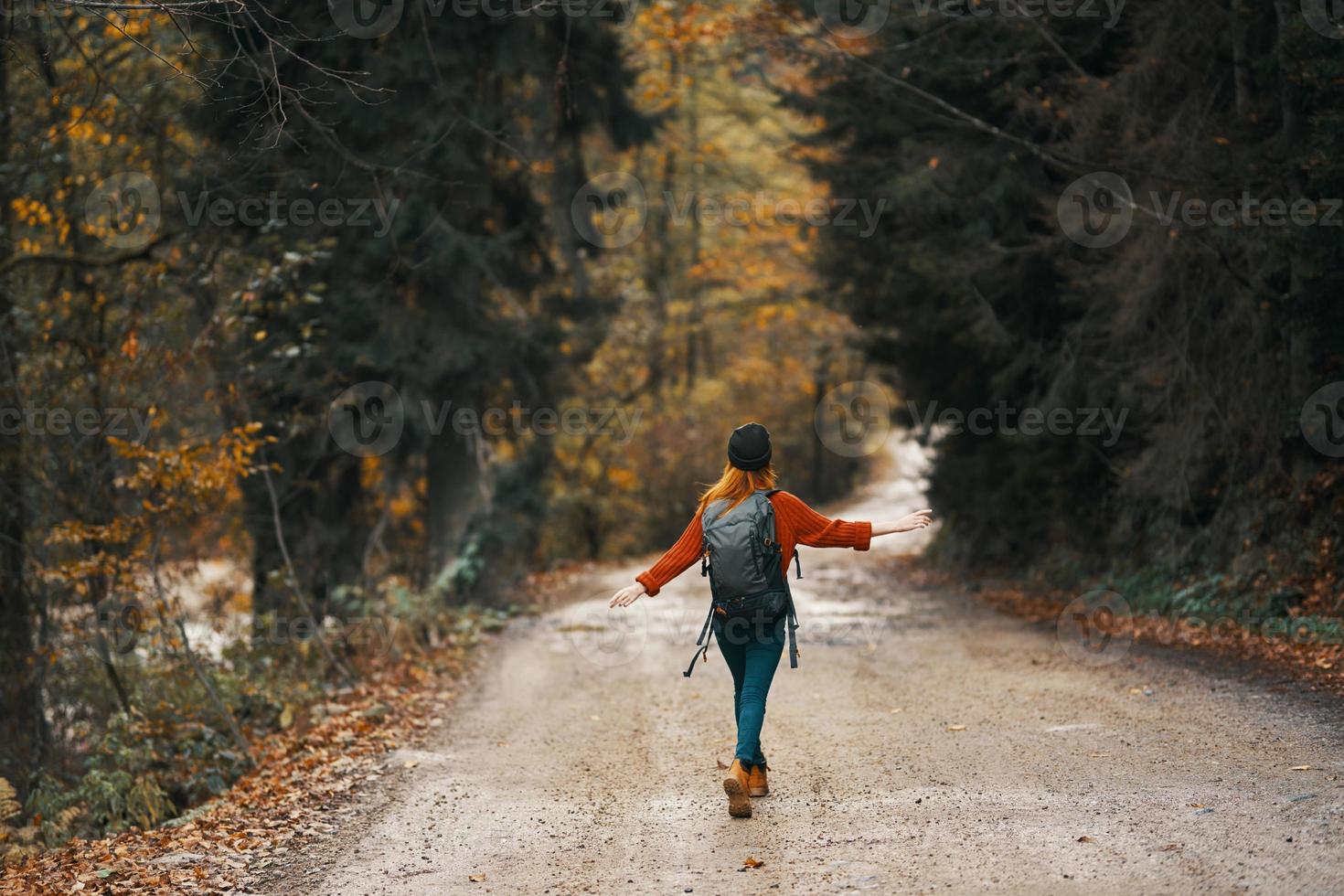 Frau im Jeans Jacke mit ein Rucksack Spaziergänge entlang das Straße im das Herbst Wald foto